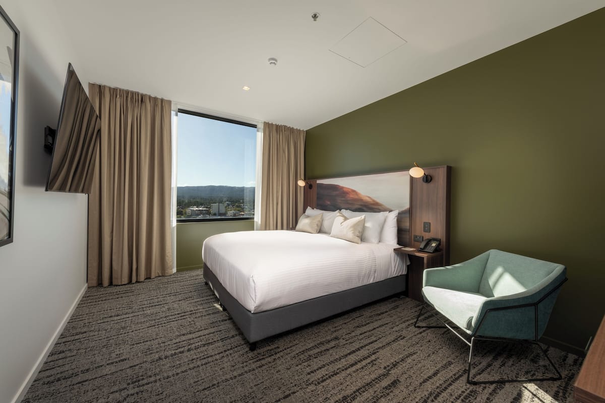 A hotel room with a large bed, a chair, a nightstand, and a view of the city.
