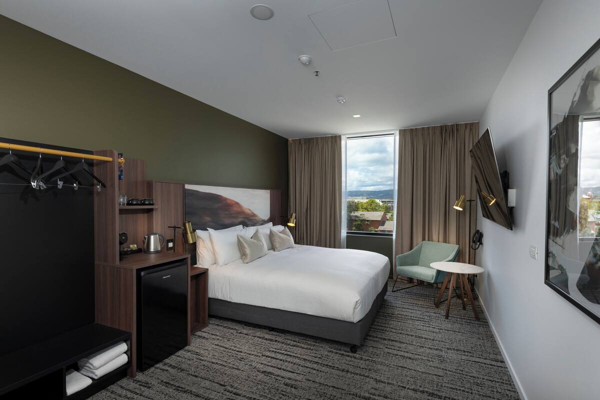 A modern hotel room with a large bed, a desk with a computer, a small seating area, and a view of the outdoors.