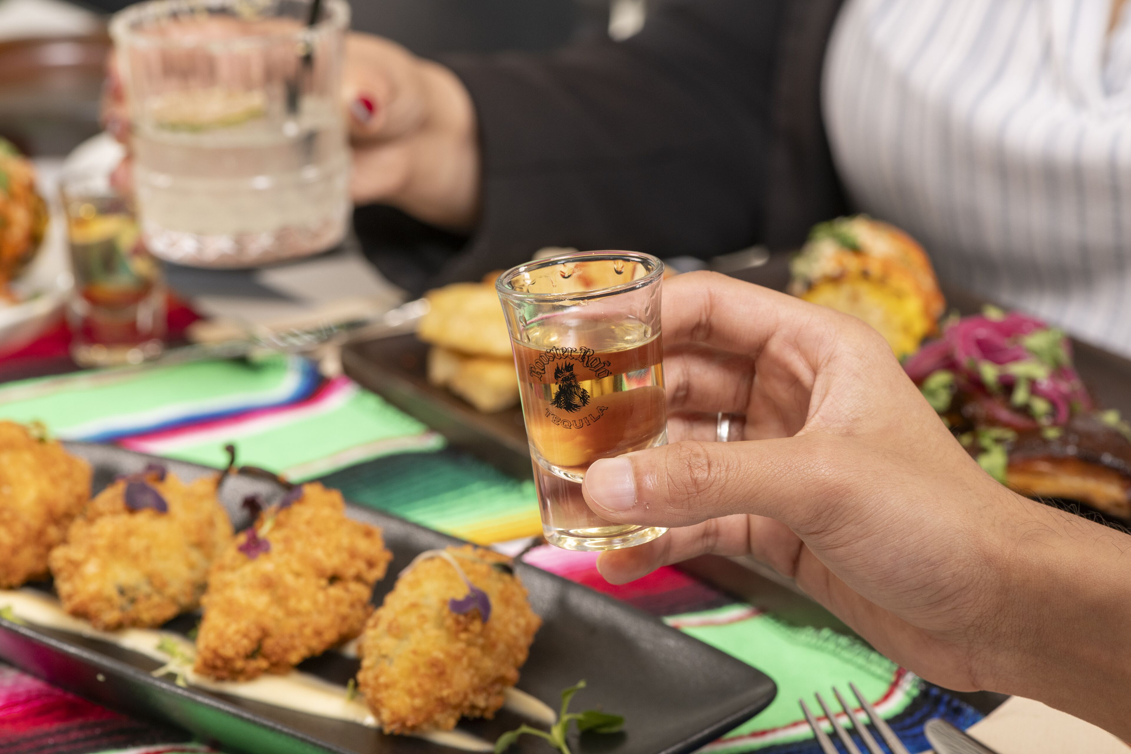 A person holding a small glass with a beverage, a plate with fried food, and a colorful tablecloth.