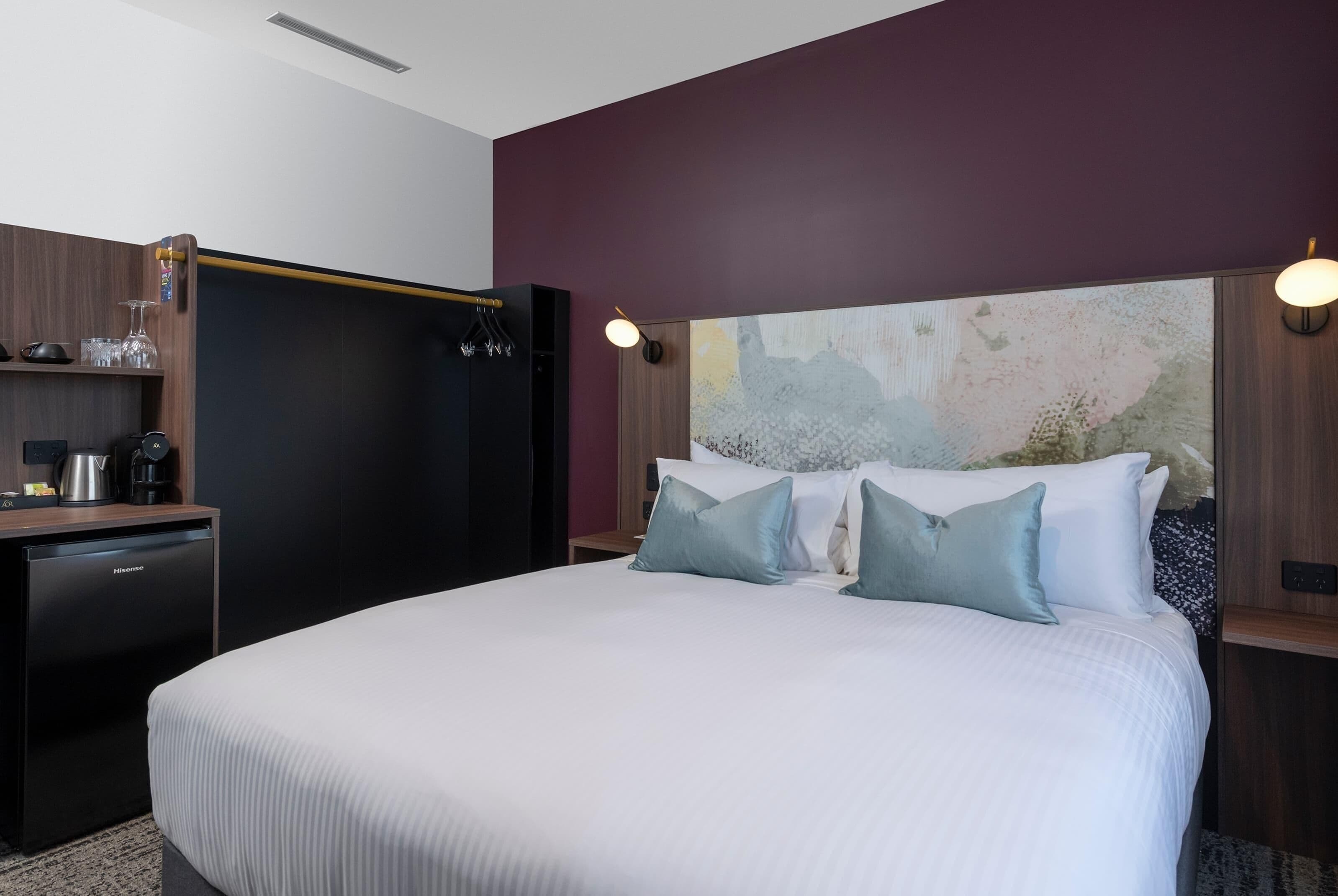 A hotel room with a large bed, a desk with a coffee maker, and a wardrobe.