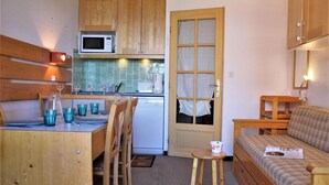 Cocina compacta con armarios de madera, microondas y tostadora. Hay una zona de comedor con mesa y sillas, y un taburete pequeño. Un banco de madera con cojín en la esquina.