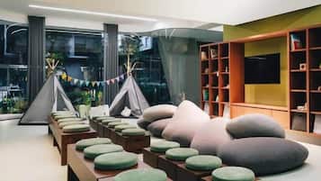 A modern living room with a long wooden table, green and gray bean bag chairs, a bookshelf, and a large window with curtains.