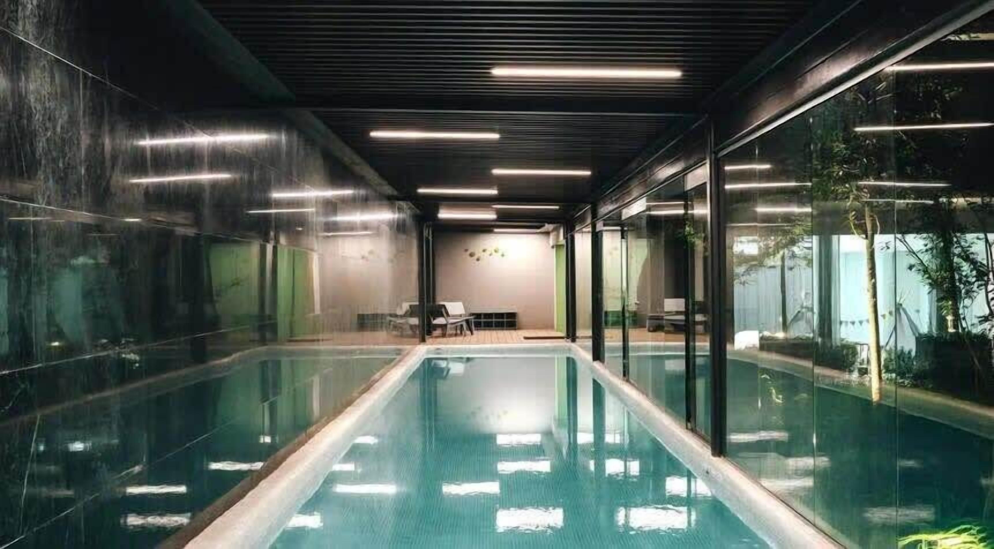 A modern indoor swimming pool with clear glass walls, a minimalist design, and a seating area with a table and chairs.