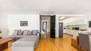 A modern living room with a sectional sofa, wooden flooring, and a kitchen area visible through an open door. - Spacious Modern Home in iconic Green Point (Cape Town)