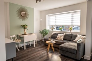 Een moderne woonkamer met een grijze bank, een eettafel en een groot raam met zonwering.