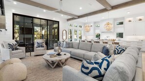 Un salon moderne avec un grand canapé, deux fauteuils, une table basse et un coin repas, la cuisine étant visible au fond.