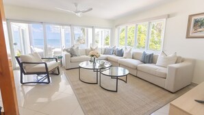 Ein geräumiges Wohnzimmer mit einem weißen Sofa, zwei runden Glastischen und einem schwarzen Sessel. Große Fenster bieten Blick auf den Ozean.