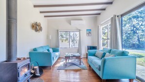 Ein Wohnzimmer mit zwei blauen Sofas, einem Glastisch, einem Kamin und großen Fenstern mit Blick ins Freie.