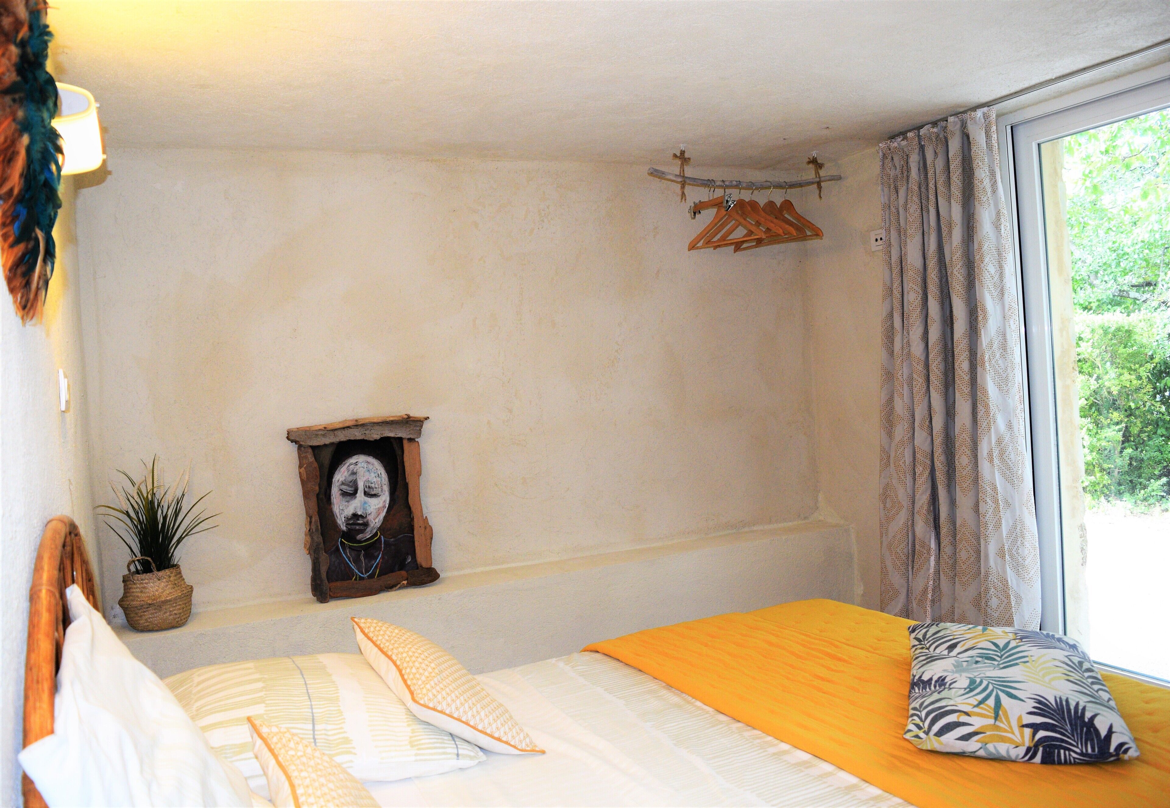 Un dormitorio con cama, ventana con cortinas, perchero de pared, una planta pequeña y un cuadro enmarcado.