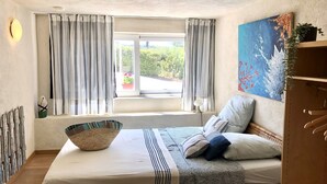 Een slaapkamer met een bed, een raam met gordijnen, een schilderij aan de muur en een plant op een plank.