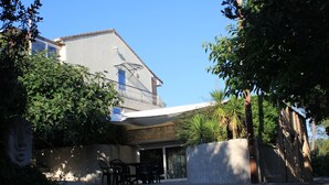 Een twee verdiepingen tellend huis met een schuin dak, een overdekte patio met tuinmeubilair en een grote boom op de voorgrond.