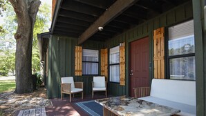 A covered porch with a wooden table and two white chairs, a red door, and a view of the outdoors.