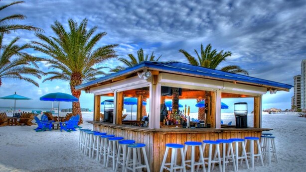 A beach bar with wooden structure, blue bar stools, and umbrellas.
