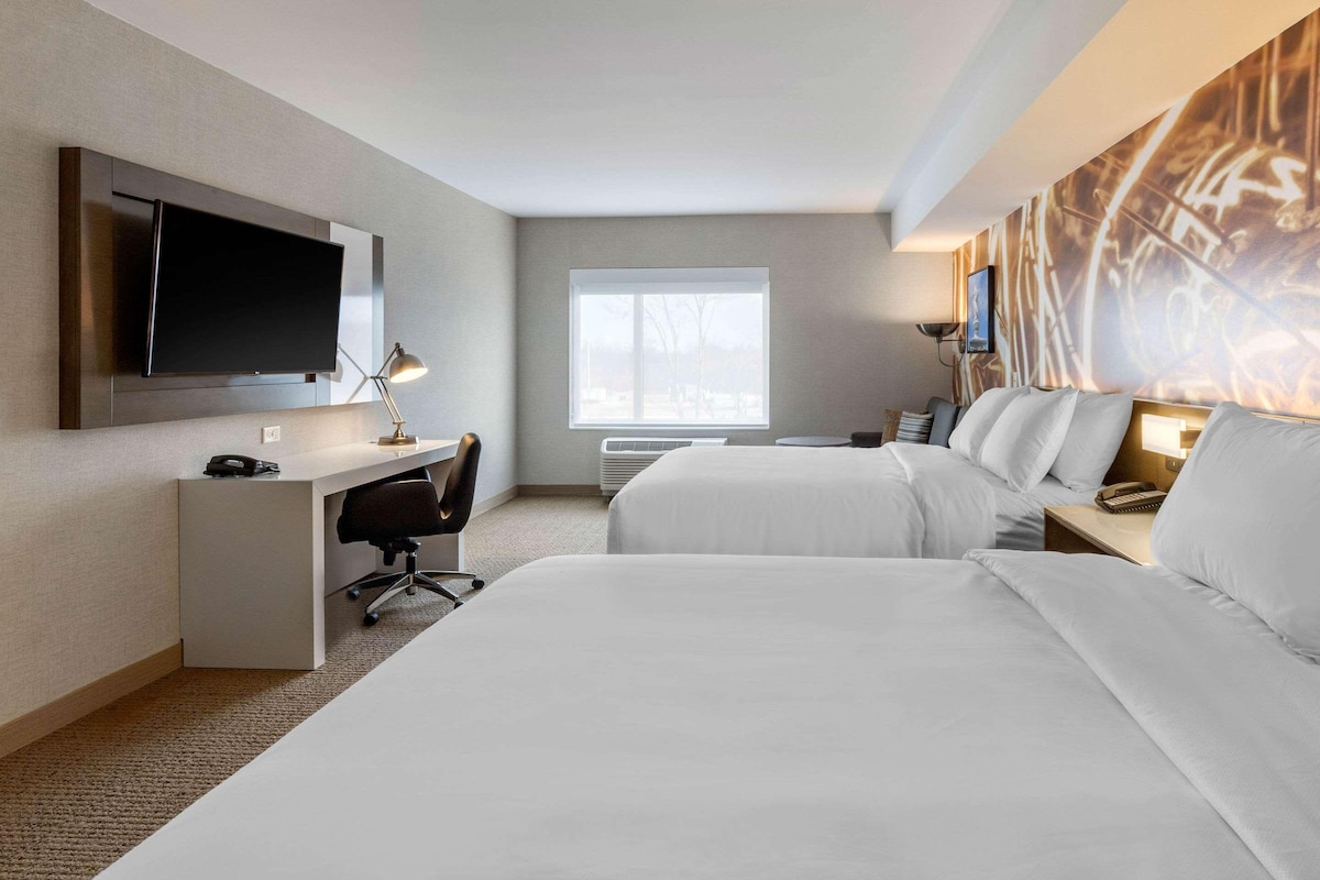 Habitación de hotel con dos camas, un escritorio con silla, un televisor de pantalla plana en la pared, una ventana y una lámpara de pared.