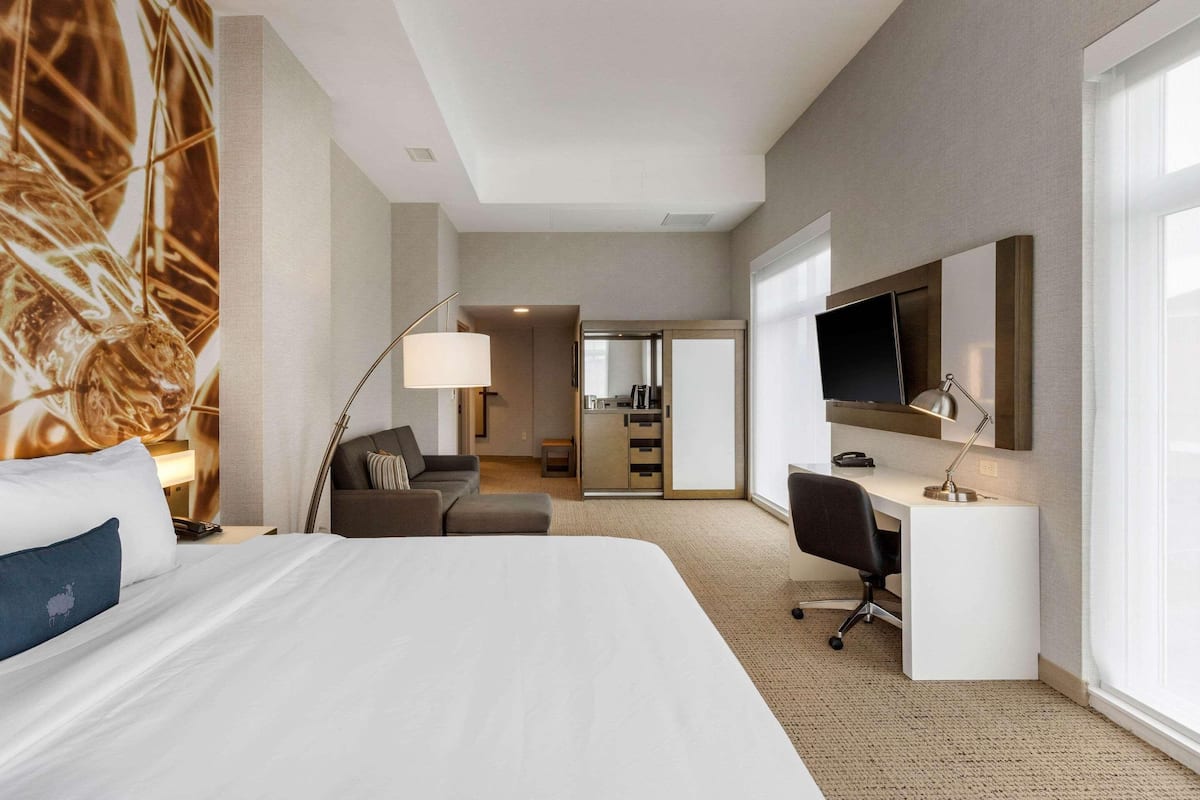 Quarto de hotel com uma cama grande, uma escrivaninha, uma cadeira, uma televisão e uma kitchenette ao fundo.