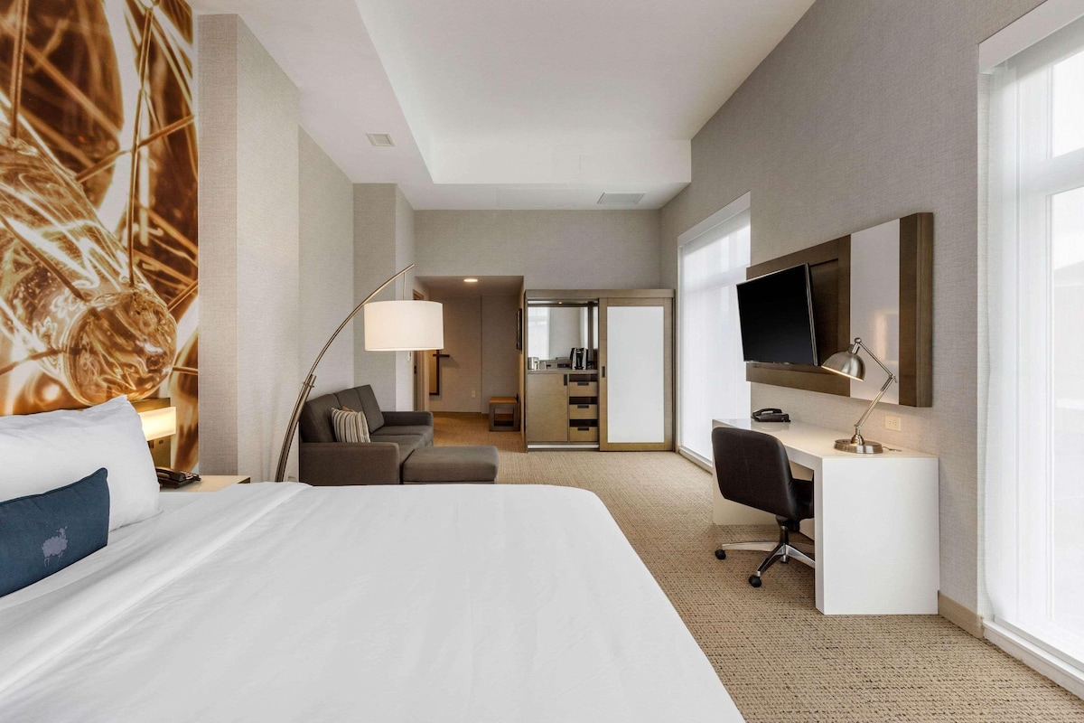 Habitación de hotel con una cama grande, un escritorio, una silla, un televisor y una kitchenette al fondo.
