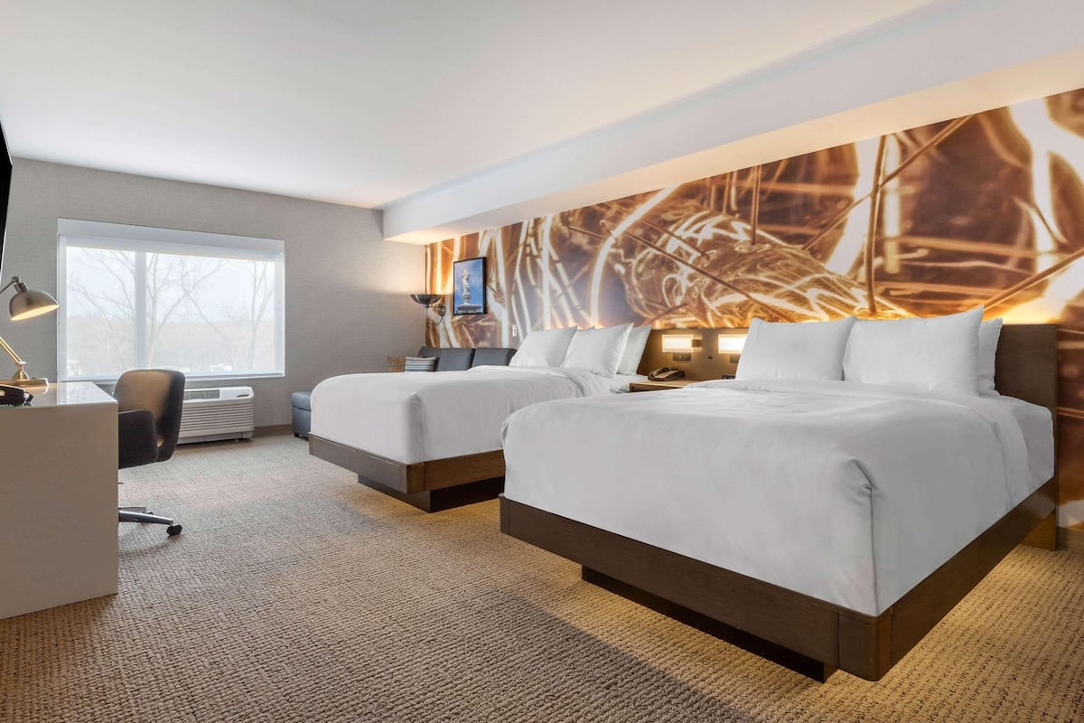 Quarto de hotel com duas camas, uma escrivaninha, uma cadeira, uma janela grande e uma parede com padrão geométrico.