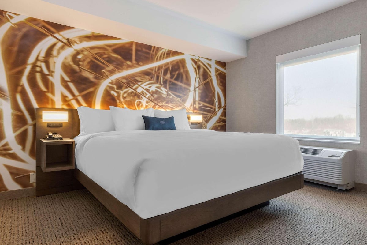 Quarto de hotel com uma cama grande, mesa de cabeceira com abajur, ar condicionado fixado na parede e uma grande janela com vista.