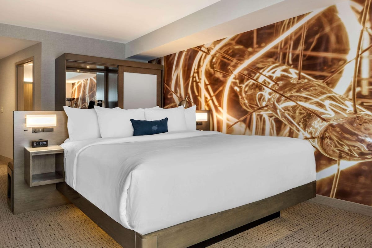 Quarto de hotel com uma cama grande, uma mesa de cabeceira com relógio, um espelho e uma parede com um mural abstrato.