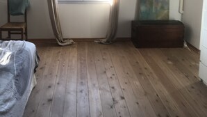 Ein Schlafzimmer mit Holzboden, einem Fenster mit Vorhängen, einem Bett, einem Sessel und einem Bild an der Wand.