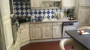 Eine Küche mit weißen Schränken, einer blau-weißen Schachbrett-Spritzschutzleiste und einem roten Boden. Ein Barhocker und ein Spüle mit verschiedenen Flaschen und Behältern auf der Arbeitsfläche sind ebenfalls vorhanden.