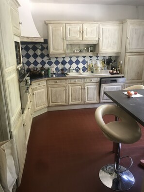 Eine Küche mit weißen Schränken, einer blau-weißen Schachbrett-Spritzschutzleiste und einem roten Boden. Ein Barhocker und ein Spüle mit verschiedenen Flaschen und Behältern auf der Arbeitsfläche sind ebenfalls vorhanden.