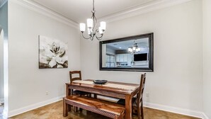 Comedor con mesa y bancos de madera, una araña, un cuadro enmarco y un espejo que refleja una cocina.