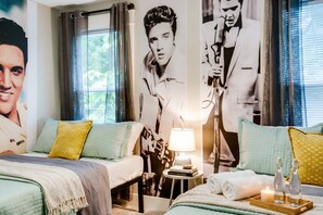 A hotel room with two beds, a bedside table with a lamp, a tray with bottles and a candle, and large posters on the walls.