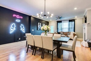 Ein modernes Wohnzimmer mit Essbereich, ausgestattet mit einer schwarzen Wand, Neonreklameschildern, einem Kronleuchter und einem Deckenventilator.