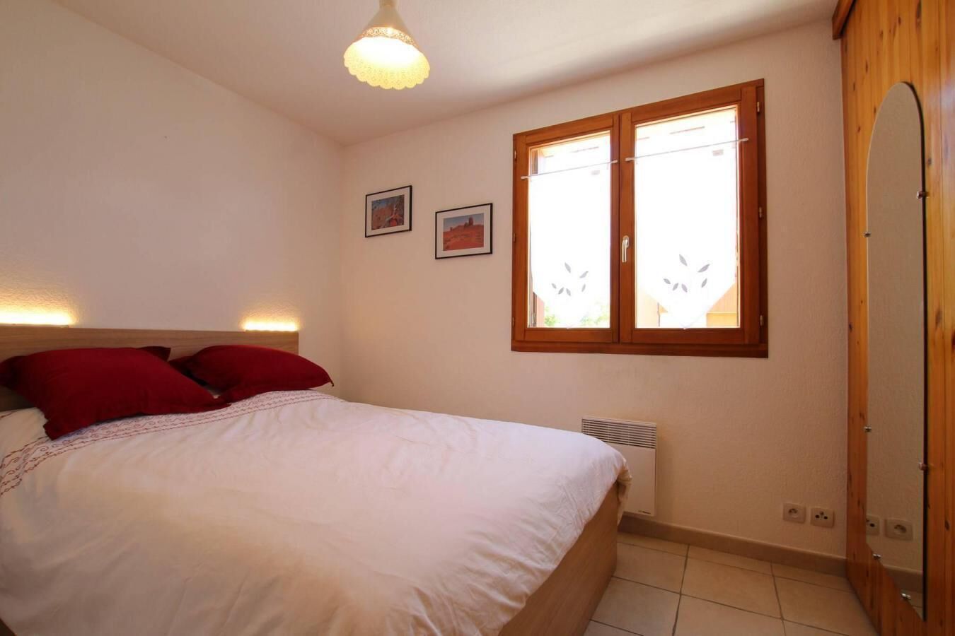 A bedroom with a bed, a window with blinds, a wall-mounted heater, and a wooden wardrobe.