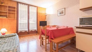 Une pièce chaleureuse avec un plancher en bois, une table à manger recouverte d’une nappe rouge, un banc, un téléviseur et un coin cuisine.