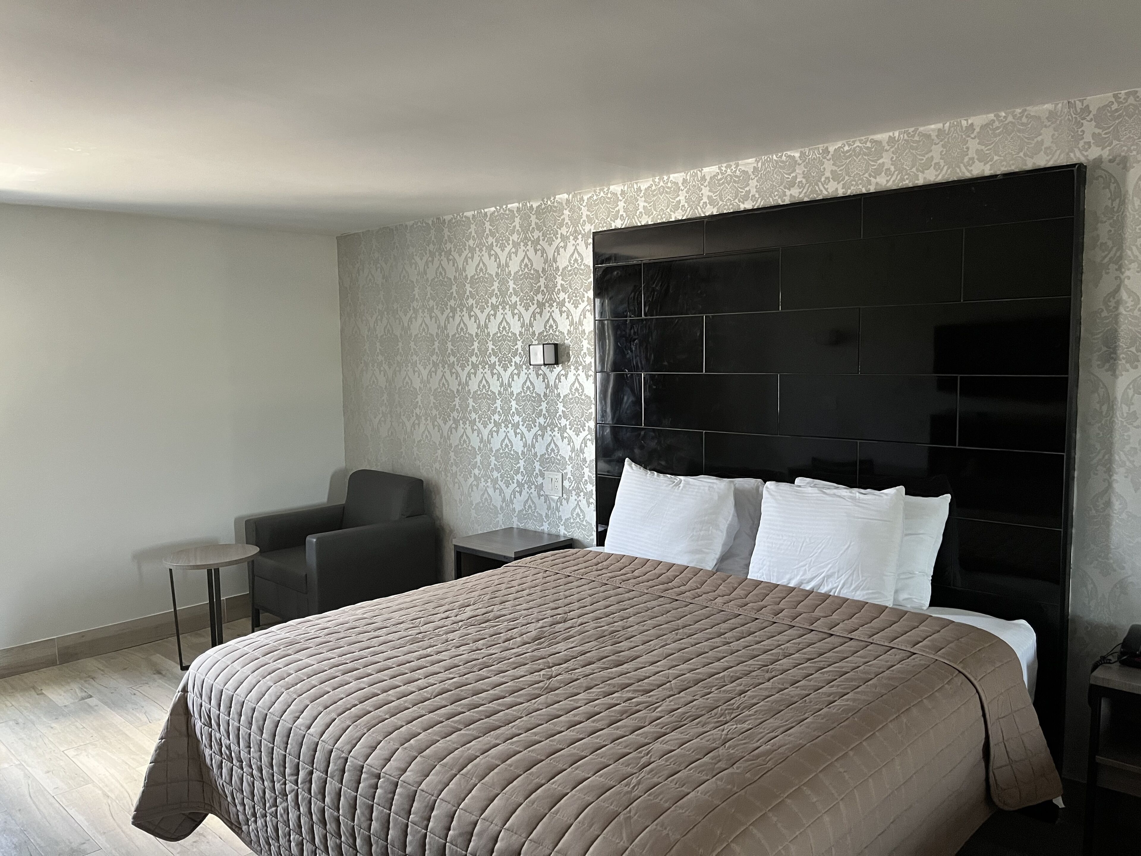 A modern hotel room with a large bed, a small round table, a chair, and a black headboard.