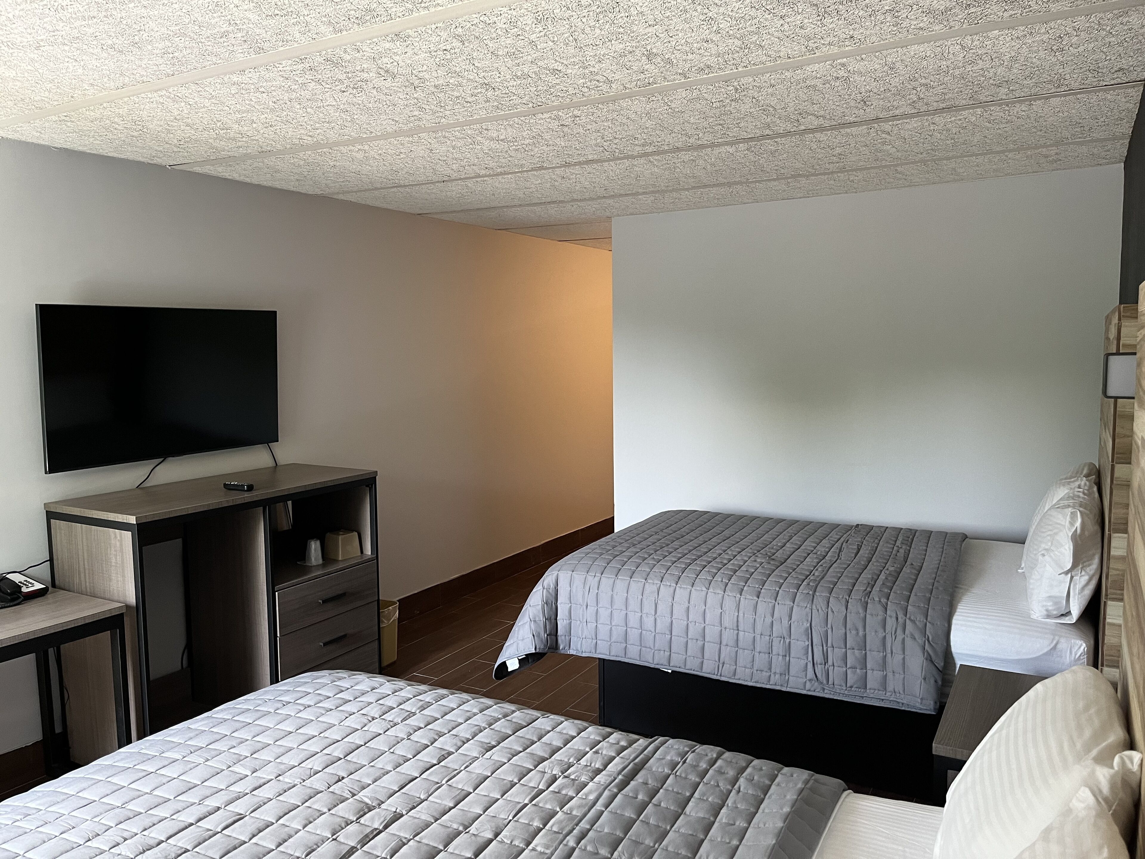 A hotel room with a flat-screen TV, a desk with a chair, and two beds with grey bedding.
