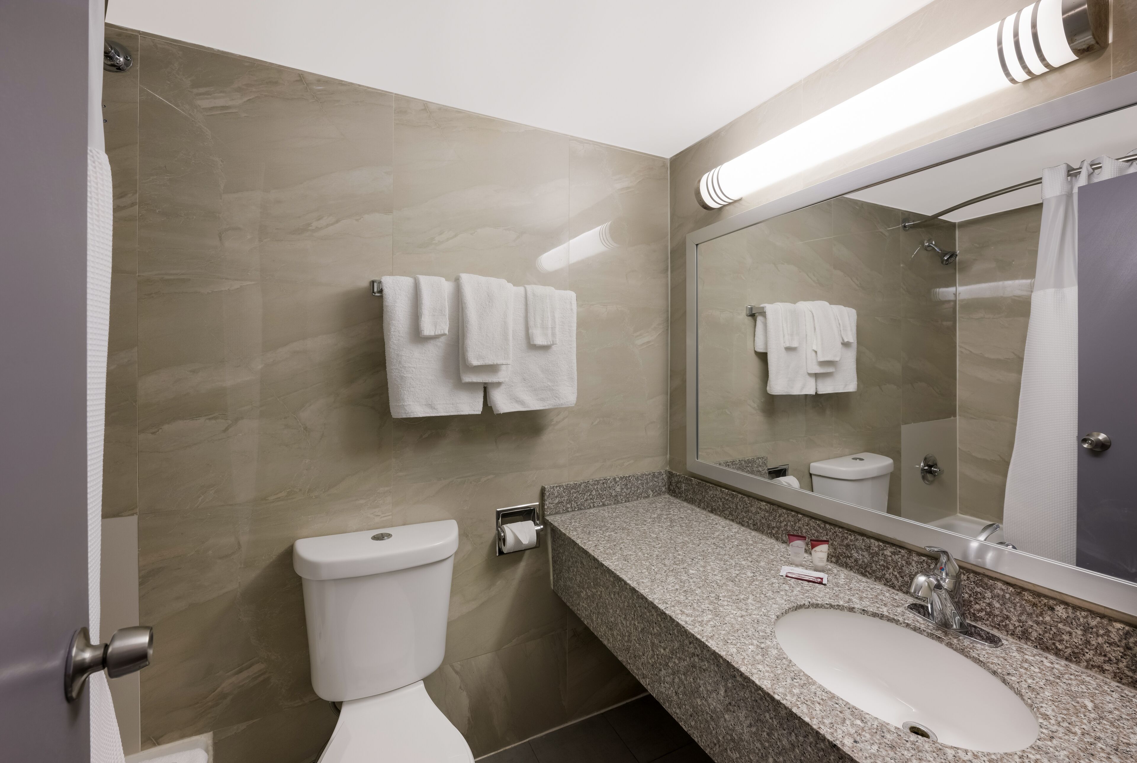 A modern bathroom with a marble wall, a white toilet, a granite countertop, a sink, and a large mirror.