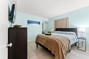 Een slaapkamer met een bed, een dressoir, een televisie, een schilderij, een lamp en een raam met gordijnen.