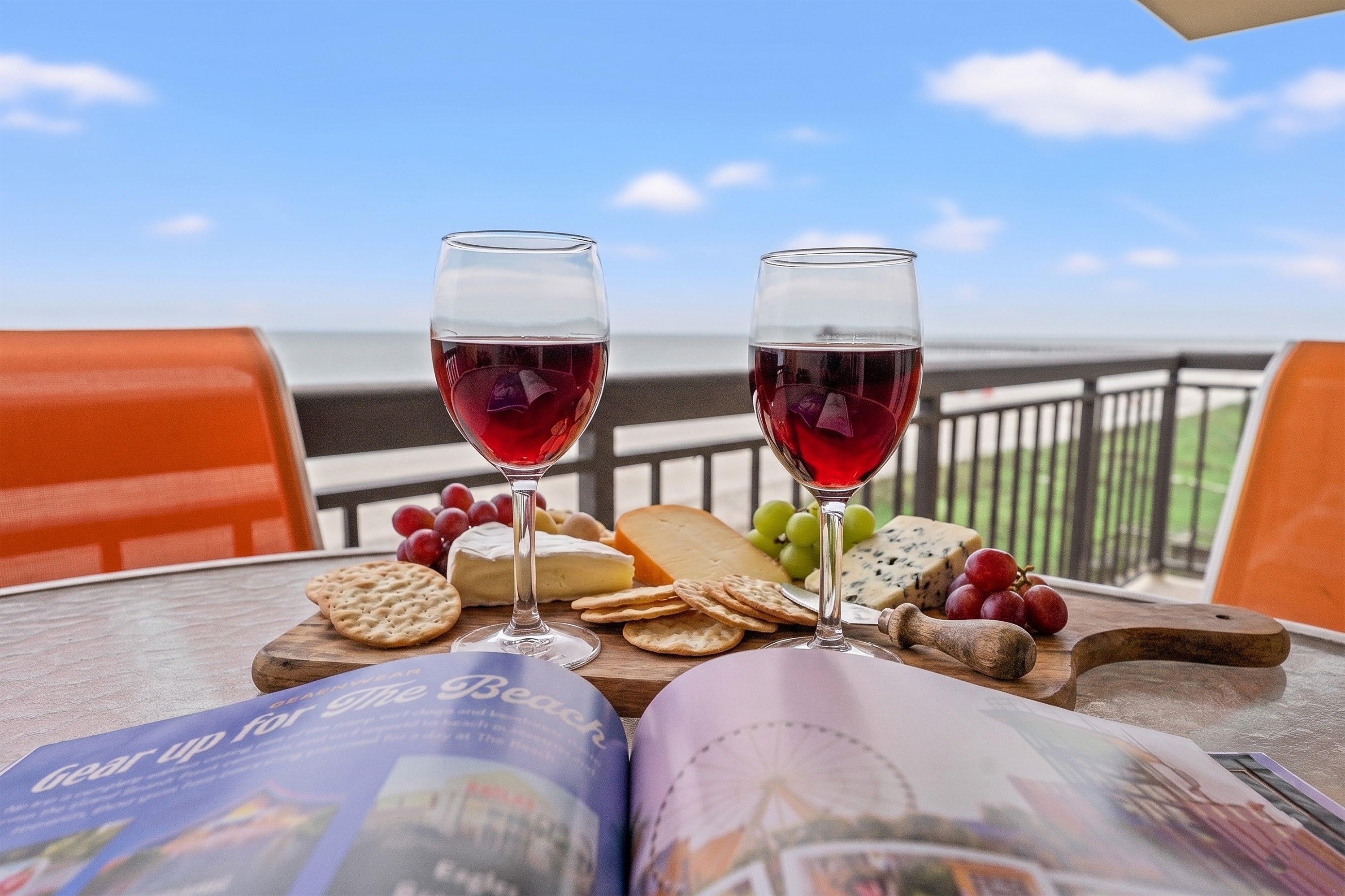 Ein Tisch mit zwei Gläsern Rotwein, einer Käseplatte mit verschiedenen Käsesorten, Crackern und Trauben, sowie einer Zeitschrift, die eine Strandansicht zeigt.
