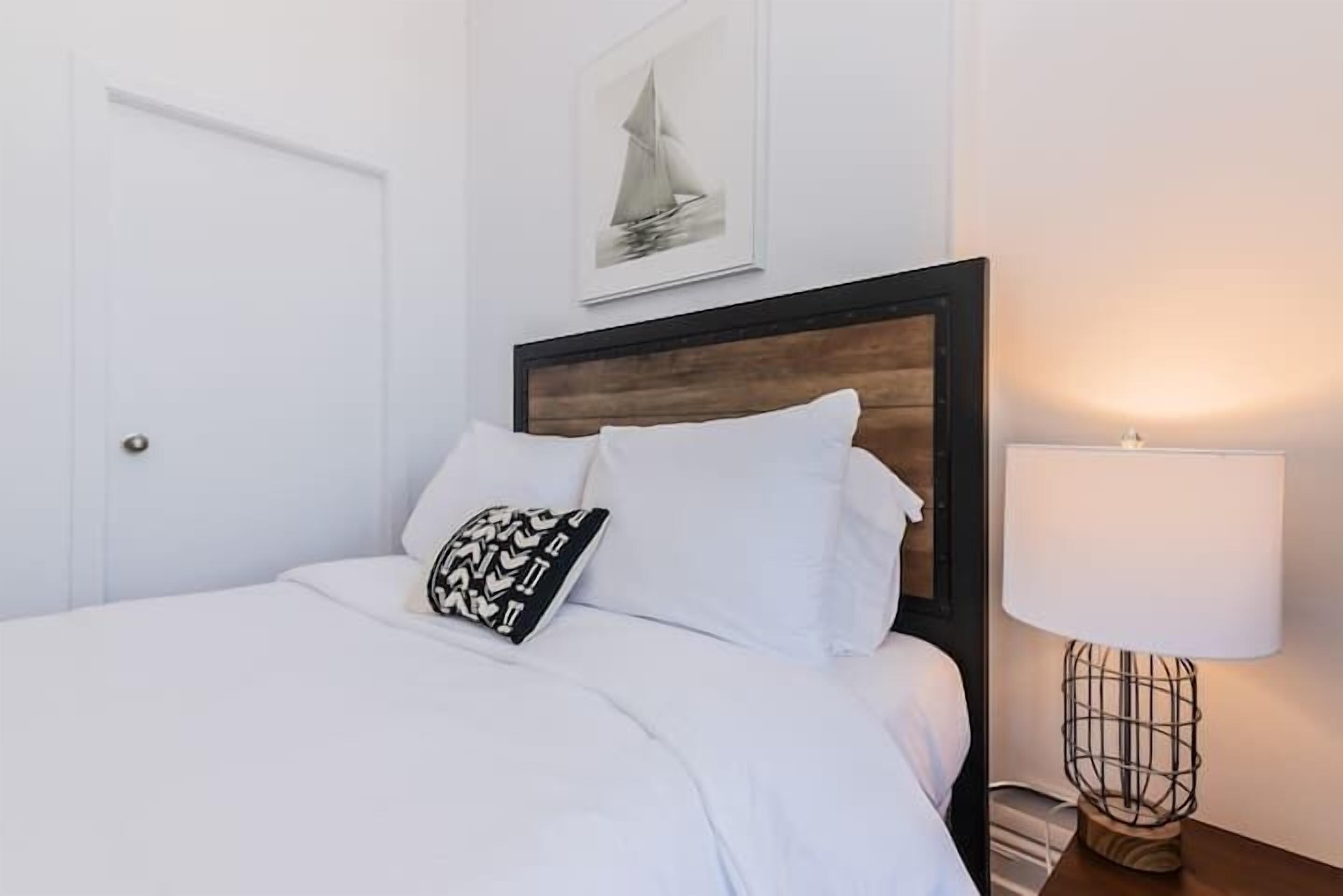Una cama bien hecha con sábanas blancas, un cojín estampado en blanco y negro, y un cabecero de madera. Encima de la cama hay un cuadro enmarcado, y una lámpara de noche se ve a la derecha.