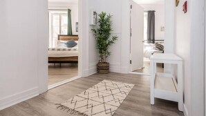 A hallway with a wooden floor, a patterned rug, a potted plant, and a white bedside table. - Manoir des Remparts 3 Bdrm Oasis (Québec City)