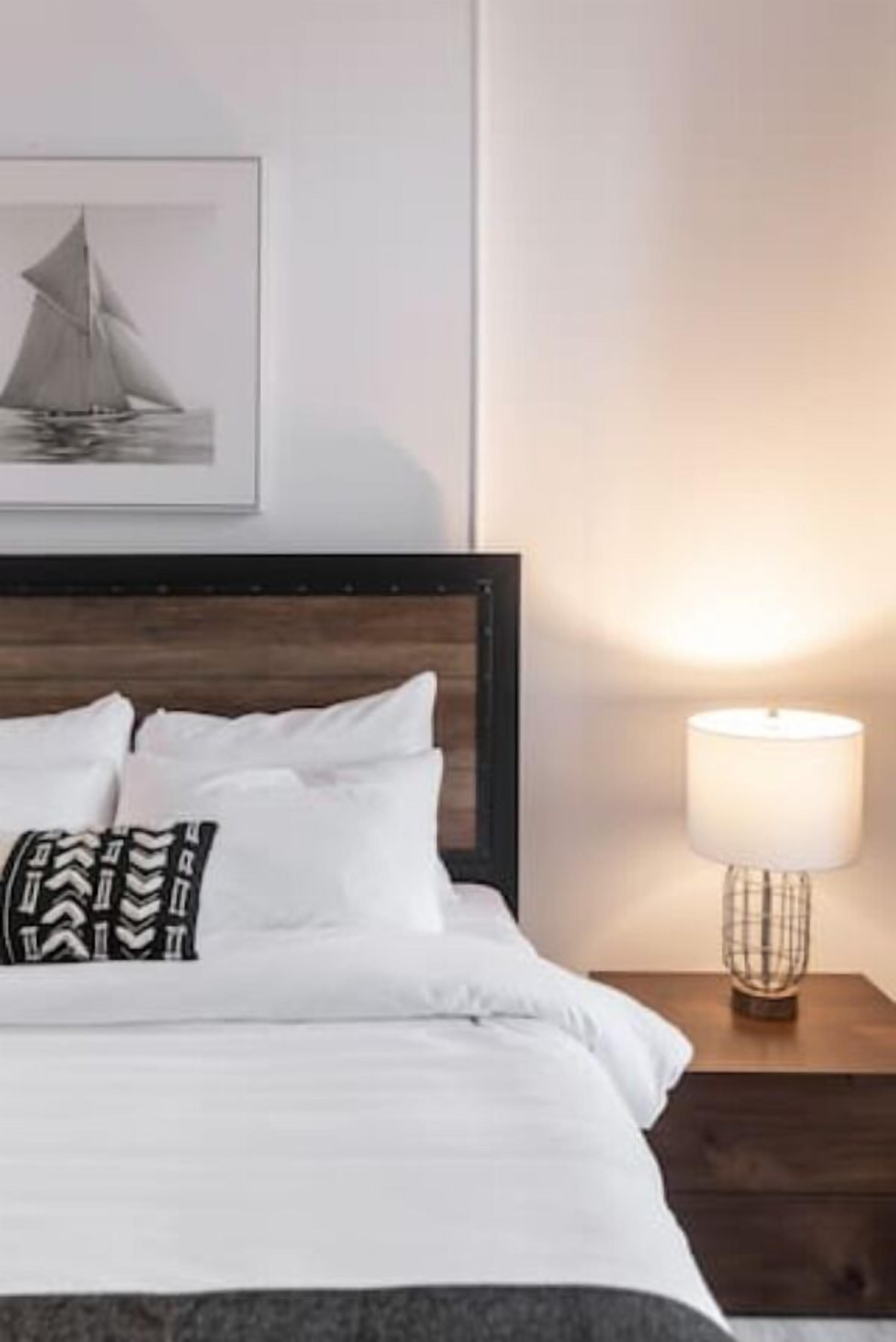 A neatly made bed with white linens and a patterned pillow, a framed picture of a sailboat on the wall, and a bedside table with a lamp.