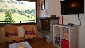 A cozy living room with a stone fireplace, wicker chairs, a flat-screen TV, and a large window overlooking a scenic landscape. - 1st floor, view ski slopes, balcony, fireplace or stove, tv, 45m², Le Grand Bornand (Le Grand-Bornand)