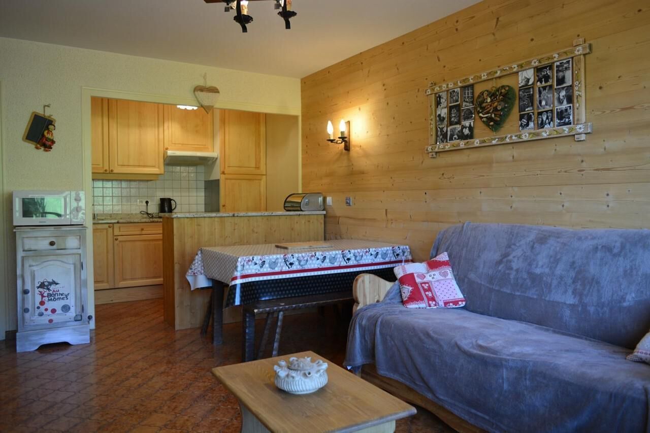 Un salon chaleureux avec un canapé et une table, des meubles en bois, un coin cuisine équipé d’un grille-pain, et un mur décoré de photos.