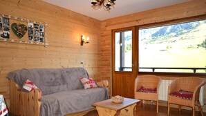 A cozy room with a wooden interior, a grey sofa, wicker chairs, a small table, and a large window offering a view of the outdoors. - 1st floor, view ski slopes, balcony, fireplace or stove, tv, 45m², Le Grand Bornand (Le Grand-Bornand)