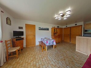 A room with a wooden floor, a table with a tablecloth, a television on a stand, wooden cabinets, and a ceiling light fixture.