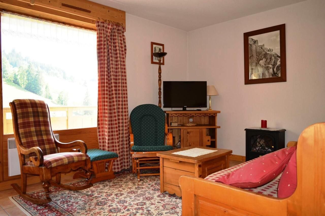 A cozy room with a rocking chair, a green armchair, a wooden desk, a television, and a fireplace.