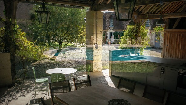 Ein überdachter Essbereich im Freien mit Pool, steinernen Säulen und Blick auf ein Haus im Hintergrund.