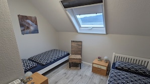 A dorm-style room with two beds, a small chair, a nightstand, and a skylight.