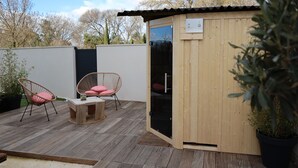 Een houten terras met twee rieten stoelen en een kleine tafel, een houten sauna en een potplant.