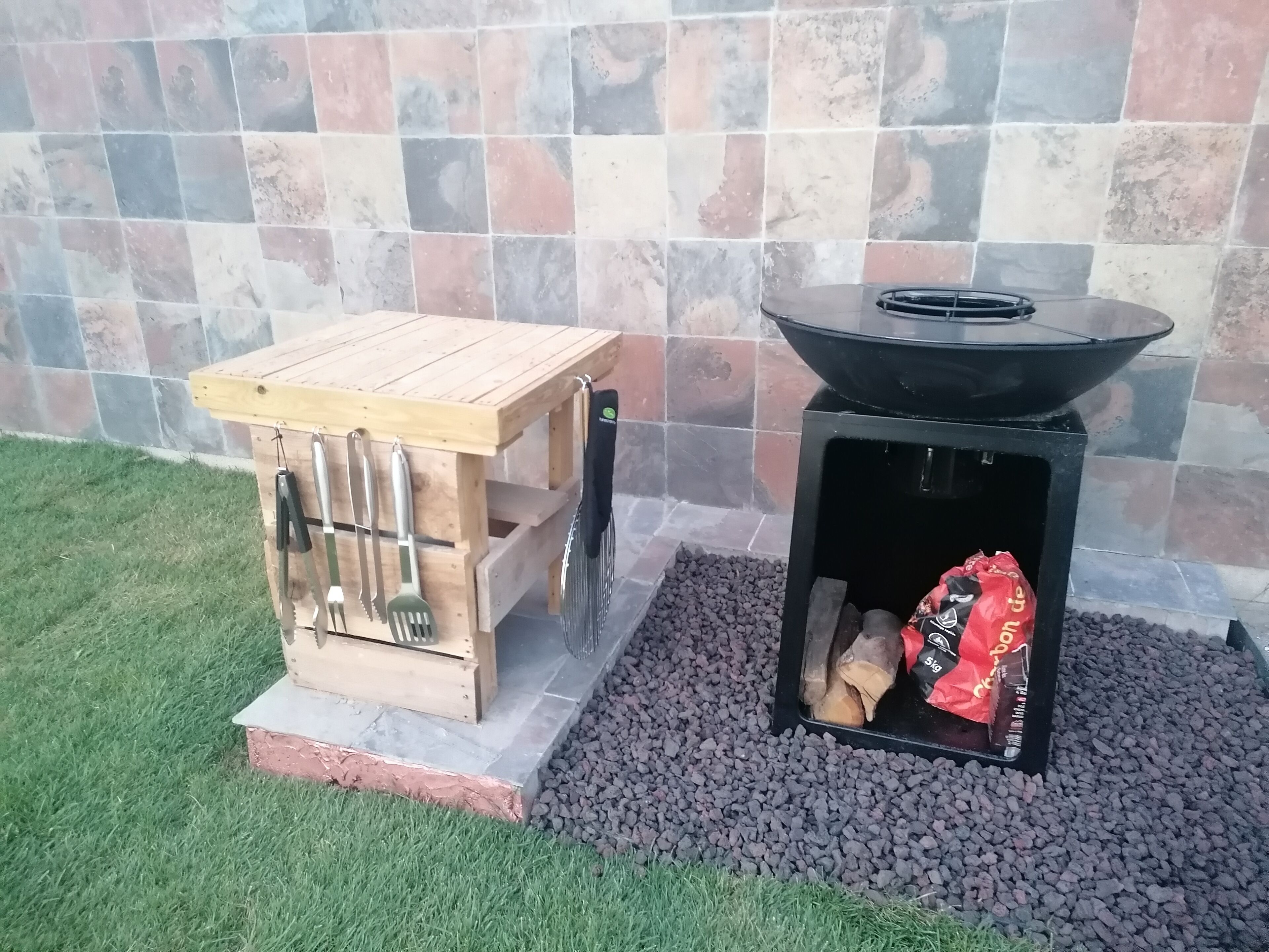 A wooden outdoor grill table with utensils and a black grill with wood and a bag of charcoal.