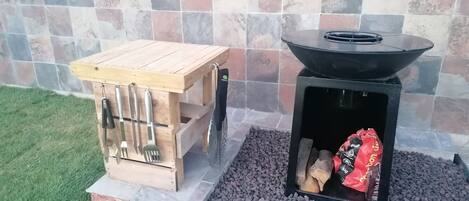 A wooden outdoor grill table with utensils and a black grill with wood and a bag of charcoal.