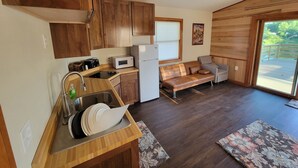 A compact kitchen with wooden cabinets, a microwave, and a sink. There's a sofa and a window with blinds. - Quaint Lakeview Cabin  (Wadena)